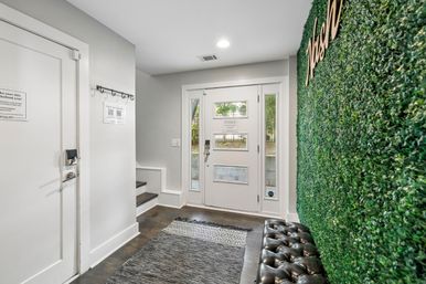 Modern entryway with white front door and sidelights, faux boxwood green accent wall, tufted black leather bench, dark hardwood floors and patterned runner rug.