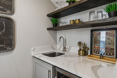 Modern home wet bar with white marble countertop and undermount sink, chrome gooseneck faucet, white herringbone tile backsplash, dark floating wood shelves with glassware, potted plants, brass accents and a framed vintage bourbon print.