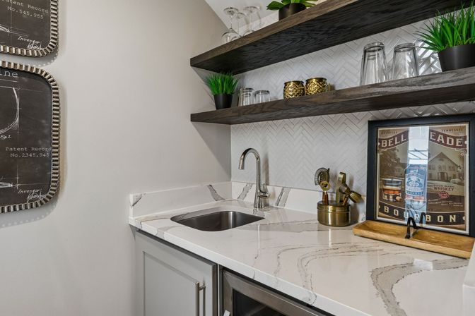Modern home wet bar with white marble countertop and undermount sink, chrome gooseneck faucet, white herringbone tile backsplash, dark floating wood shelves with glassware, potted plants, brass accents and a framed vintage bourbon print.
