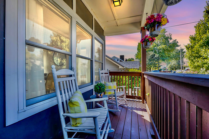 Cozy front porch with two white rocking chairs with yellow-striped cushions, small side table with a potted plant, hanging flower baskets and wooden railing overlooking a leafy suburban street at sunset.