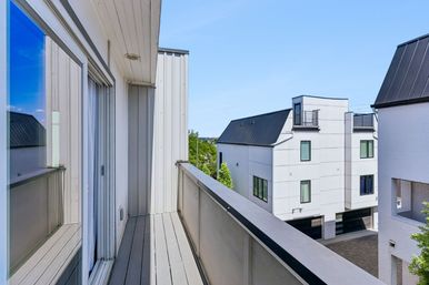 Sunny narrow balcony with sliding glass door and composite decking overlooking modern white townhouses with black metal roofs and a small courtyard under a clear blue sky.