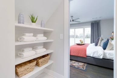 Built-in linen shelves with neatly folded white towels, wicker baskets, striped vases and a small potted plant, opening into a bright modern master bedroom with a coral quilt, layered pillows, tray of flowers and sliding glass doors.