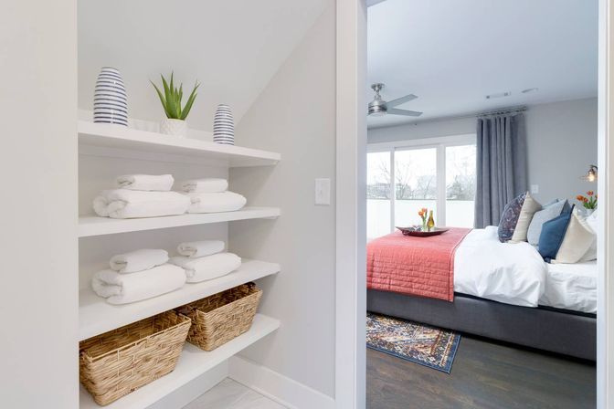Built-in linen shelves with neatly folded white towels, wicker baskets, striped vases and a small potted plant, opening into a bright modern master bedroom with a coral quilt, layered pillows, tray of flowers and sliding glass doors.