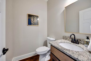 Cozy modern powder room with granite vanity countertop, dark bronze faucet, white toilet, wood flooring, neutral beige walls, and framed gold-leaf botanical artwork.