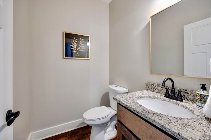 Cozy modern powder room with granite vanity countertop, dark bronze faucet, white toilet, wood flooring, neutral beige walls, and framed gold-leaf botanical artwork.