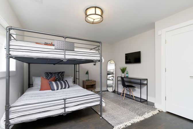 Bright modern bedroom with industrial metal bunk bed, striped bedding, study desk with wall-mounted TV, full-length mirror and woven rug.