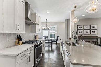 Bright modern open-concept kitchen with white shaker cabinets, stainless steel range and hood, herringbone tile backsplash, large quartz island with undermount sink, glass pendant lights, dark hardwood floors and a cozy dining nook