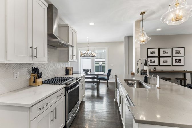Bright modern open-concept kitchen with white shaker cabinets, stainless steel range and hood, herringbone tile backsplash, large quartz island with undermount sink, glass pendant lights, dark hardwood floors and a cozy dining nook