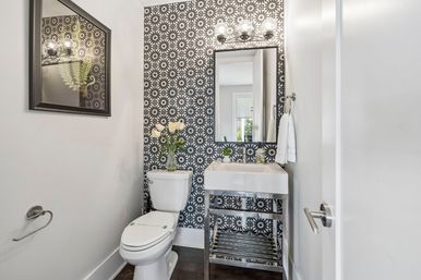 Small modern powder room with bold black-and-white floral wallpaper, rectangular mirror and globe light, white toilet topped with a vase of tulips, and a white sink on a stainless-steel open-frame vanity