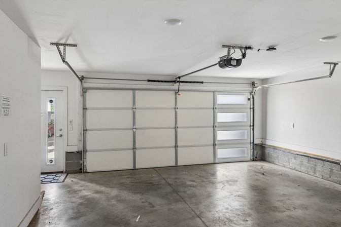Empty two-car residential garage with concrete floor, white sectional garage door featuring frosted windows, ceiling-mounted opener and side entry door.