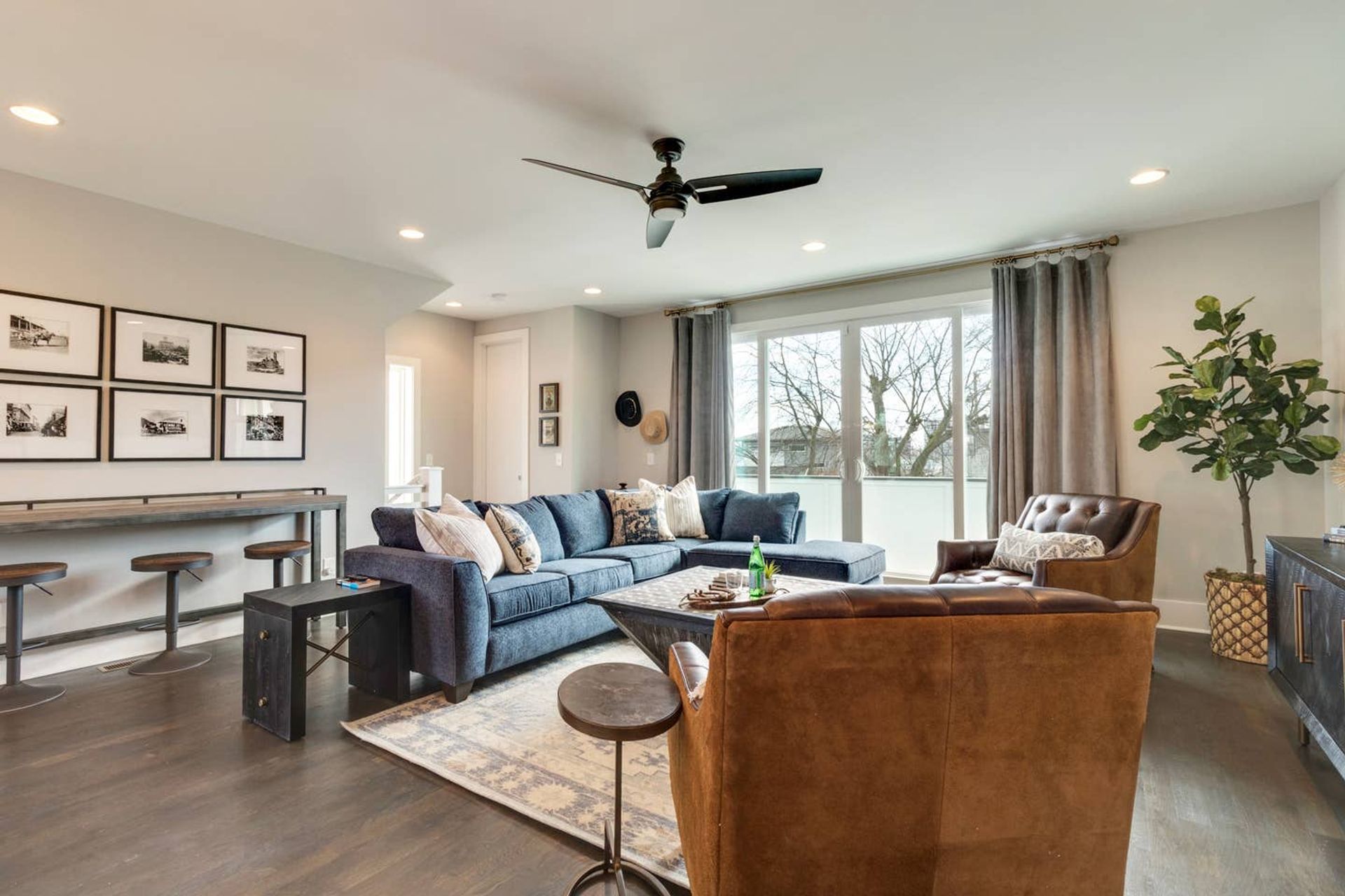 Bright modern living room with blue sectional sofa, brown leather armchairs, dark hardwood floors, ceiling fan, large sliding glass doors to a balcony, framed gallery wall and tall potted plant