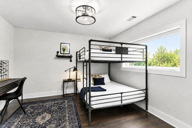 Bright modern bedroom with black metal bunk bed, large horizontal window with green tree view, dark hardwood floors, navy patterned rug, small desk and industrial ceiling light