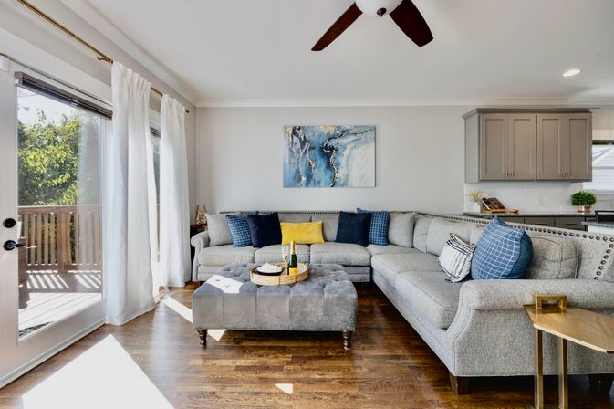 Sunlit modern living room with gray L-shaped sectional and nailhead trim, tufted ottoman with serving tray and champagne, blue and yellow throw pillows, abstract blue wall art, hardwood floors and sliding glass doors to a wooden deck.