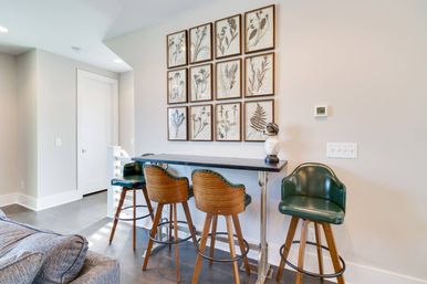 Cozy modern home bar nook in a light living area with four green leather swivel bar stools on wooden legs, a black counter table, a 3x4 grid of framed botanical prints on the wall, and hardwood floors.