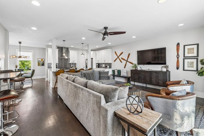 Open-concept modern living room and kitchen in a suburban home — large gray sectional, dark hardwood floors, island with pendant lights and bar stools, wall-mounted TV, aviator-style chairs and wooden propeller wall decor.
