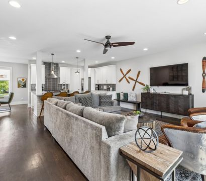 Open-concept modern living room and kitchen in a suburban home — large gray sectional, dark hardwood floors, island with pendant lights and bar stools, wall-mounted TV, aviator-style chairs and wooden propeller wall decor.