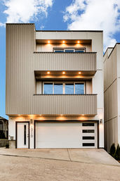 Modern three-story beige townhouse exterior with stacked balconies, warm recessed lights, white garage with horizontal windows, and a clear blue sky — urban residential home.