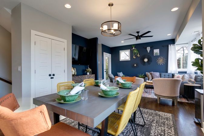 Open-concept dining and living room in a contemporary home featuring a navy accent wall, modern chandelier, gray dining table with yellow woven chairs and green place settings, cozy tufted sofa and armchairs, hardwood floors and large windows letting in natural light.