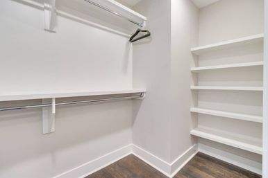 Empty modern walk-in closet with white built-in shelves, chrome double hanging rods, single black hanger and dark hardwood floor.