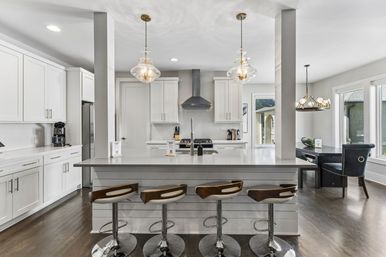 Bright modern open-plan kitchen with white cabinetry, large quartz island and breakfast bar with four wood-accent swivel stools, glass pendant lights, stainless steel range hood, dark hardwood floors and sunny dining nook by windows.