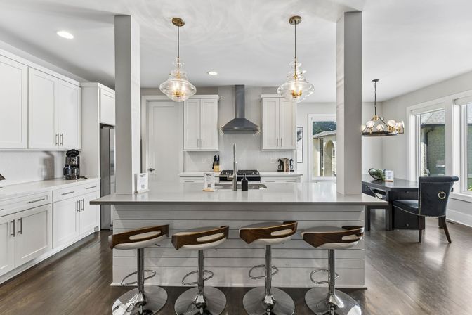 Bright modern open-plan kitchen with white cabinetry, large quartz island and breakfast bar with four wood-accent swivel stools, glass pendant lights, stainless steel range hood, dark hardwood floors and sunny dining nook by windows.