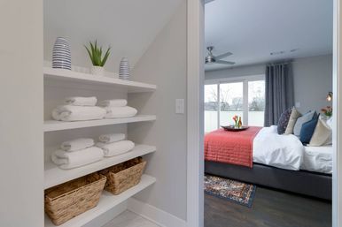 Built-in linen shelves with rolled white towels, wicker baskets and decorative vases beside a modern gray bedroom with a red quilt, layered pillows, and large windows.