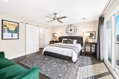Sunlit modern master bedroom with king bed and tufted gray headboard, large round wall clock, ceiling fan, sliding glass doors, green velvet sofa and patterned area rug.