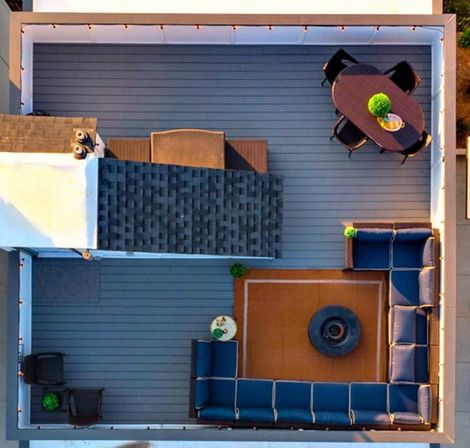 Aerial drone view of an urban rooftop deck with gray composite boards, an L-shaped navy outdoor sectional around a central fire pit on a brown rug, an oval dining table with chairs, string lights along the perimeter, and small potted plants.