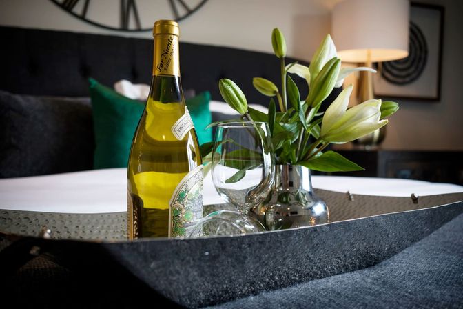 White wine bottle, two wine glasses and a vase of white lilies on a hammered metal tray atop a neatly made bed in a cozy bedroom setting.