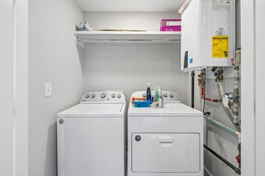 Compact bright laundry room with side-by-side white top-load washer and dryer, shelf with supplies above, iron and detergent on the dryer, and a wall-mounted tankless water heater with exposed pipes.