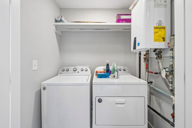 Compact bright laundry room with side-by-side white top-load washer and dryer, shelf with supplies above, iron and detergent on the dryer, and a wall-mounted tankless water heater with exposed pipes.