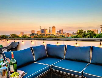 Cozy rooftop patio with navy blue cushioned sectional, hanging string lights, a bottle of wine and bowl of fruit on a table, and a glowing urban skyline at sunset.
