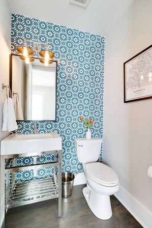 Bright modern powder room with blue geometric tile accent wall, stainless-steel vanity and white sink, rectangular mirror under two brass sconces, white toilet topped with a vase of orange tulips, framed map art and wood-look flooring.