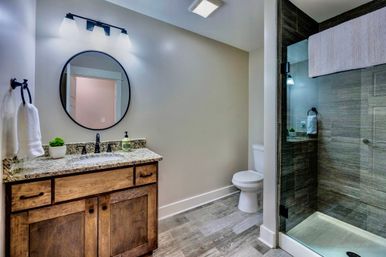 Cozy modern bathroom with rustic wood vanity and granite countertop, round black mirror, white toilet, wood-look floor and glass-enclosed gray-tiled shower with towel.