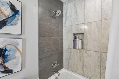 Modern bathroom bathtub and shower with gray and marble wall tiles, chrome fixtures, recessed shelf holding shampoo bottles, and abstract blue-and-gold framed artwork.