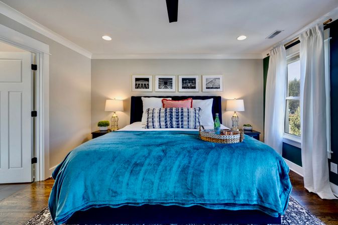 Bright contemporary master bedroom with teal blanket on king bed, patterned pillows and black upholstered headboard, bedside lamps and nightstands, framed black-and-white photos above the bed, wicker tray with a bottle, large white-curtained window and hardwood floors.
