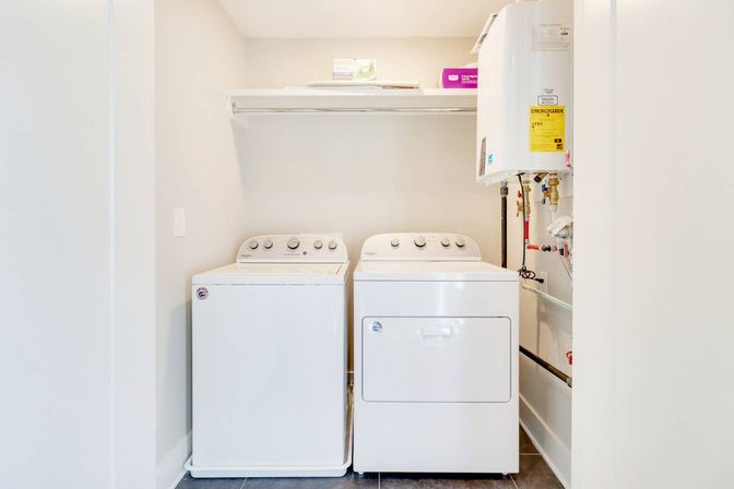 Bright compact laundry closet with white top-load washer and front-load dryer side by side under a shelf, with a water heater and visible utility hookups on the right.