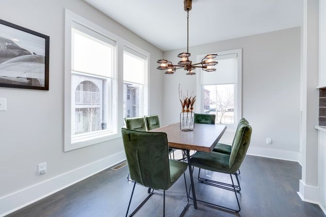 Modern, sunlit dining room with a rectangular wooden table, four green velvet chairs, an industrial-style chandelier, large windows and a vase of dried grasses