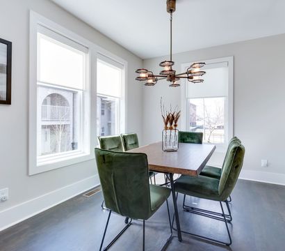 Modern, sunlit dining room with a rectangular wooden table, four green velvet chairs, an industrial-style chandelier, large windows and a vase of dried grasses