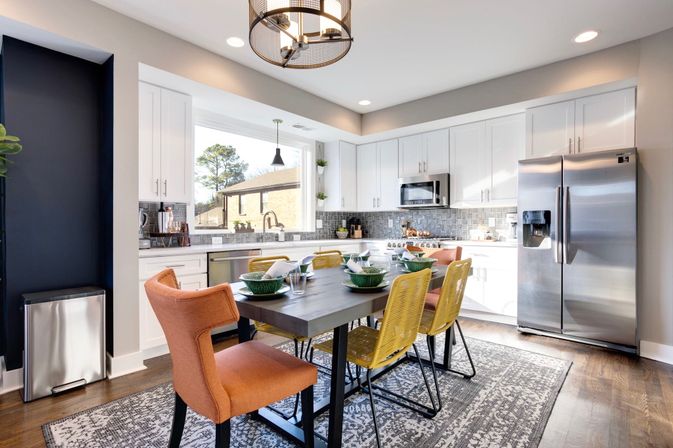 Bright modern open-concept kitchen with white cabinetry, stainless steel refrigerator and microwave, gray tile backsplash, dark wood dining table set with green bowls and cheerful orange and yellow chairs, large window and hardwood floors