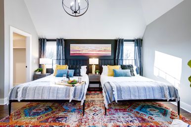 Sunlit modern twin bedroom with two beds, gray-striped throws, bright yellow and blue pillows, central nightstand and lamp, colorful patterned rug and breakfast tray.