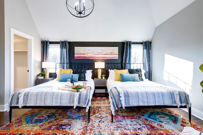 Sunlit modern twin bedroom with two beds, gray-striped throws, bright yellow and blue pillows, central nightstand and lamp, colorful patterned rug and breakfast tray.