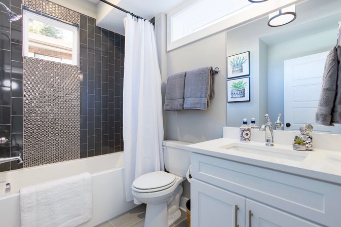 Bright modern residential bathroom with white vanity and sink, toilet, bathtub/shower with dark subway tile and mosaic accent, white shower curtain, gray towels, framed botanical prints, and chrome fixtures.