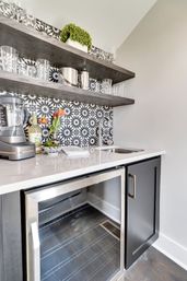 Modern home wet bar with black-and-white patterned tile backsplash, dark floating wood shelves stocked with glassware, quartz countertop with small stainless sink, blender and tulips, and a glass-front beverage fridge.