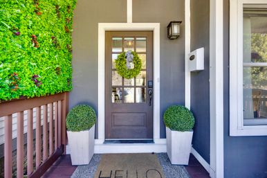 Charming gray front porch with glass-paneled brown door, green wreath, matching potted boxwoods, vertical living wall, and 'HELLO' welcome mat