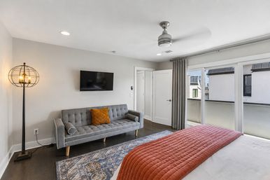 Modern bright bedroom with sliding glass doors to a small balcony, gray tufted sofa, wall-mounted TV, globe floor lamp, patterned rug and rust-colored quilted bed runner.
