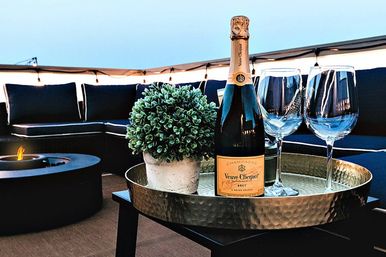 Champagne bottle and two wine glasses on a hammered tray with a potted plant on a cozy rooftop patio at dusk, fire pit and string lights in the background