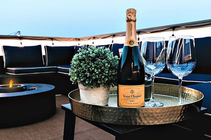Champagne bottle and two wine glasses on a hammered tray with a potted plant on a cozy rooftop patio at dusk, fire pit and string lights in the background