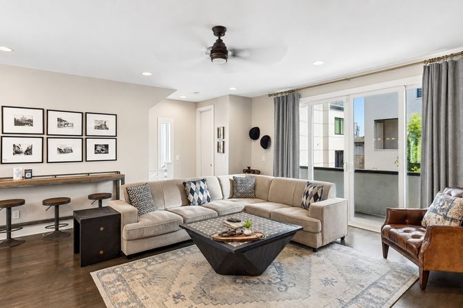 Stylish, sunlit modern living room in a city apartment with a beige L-shaped sectional and patterned pillows, geometric black coffee table on a neutral area rug, brown leather accent chair, gallery photo wall above a wood console with bar stools, sliding glass doors to a balcony, gray curtains, hardwood floors and a ceiling fan.