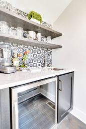 Sleek modern wet bar with black-and-white geometric tile backsplash, two floating wood shelves holding glassware and stainless barware, white countertop with flowers and blender, stainless sink and glass-front mini fridge.